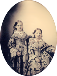 Овальный фотографический портрет в рамке двух девушек, одетых в изысканные платья викторианской эпохи.