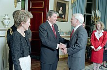White-haired man in suit greets dark-haired man in suit in formal setting, as gaunt, well-coiffed woman looks on