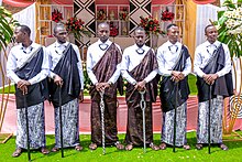 Sechs festlich gekleidete Männer mittleren Alters stehen in einer Reihe. Die Mishanana der mittleren beiden sind braun, die der äußeren vier schwarz. Die Schulterüberwürfe gehen jeweils über die linke Schulter und darunter tragen sie weiße Hemden. Sie tragen zudem Sandalen sowie dicke, schwarz-weiße Perlenketten und halten jeweils einen schwarz-weißen Spazierstock auf dem sie beide Hände ruhen. Im Hintergrund steht ein Regal mit Blumensträußen und traditionellen ruandischen Gegenständen wie zum Beispiel spezielle Körbe.