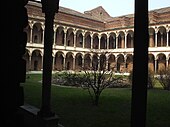 Image photographique d'un cloître. Deux colonnes au premier plan encadrent une cour verdoyante et un rond central avec au fond deux ailes à angle droit d'un édifice constitué s'arcades superposées sur deux niveaux surmontés par une toiture.