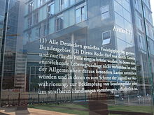 Fotografie. Freistehende Glasscheibe, mit eingraviertem Text von Artikel 11. Jakob-Kaiser-Haus sichtbar durch Scheibe. Marie-Elisabeth-Lüders-Haus spiegelt sich in Scheibe