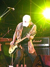 Cận cảnh một người đàn ông cúi đầu đứng trên sân khấu, tay cầm guitar, mặc quần jean và áo rộng; mặt bị chiếc mũ lưỡi trai trắng có ngôi sao đỏ che mất; phía sau anh là nhiều nhạc cụ.