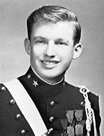 A black-and-white photograph of Donald Trump as a teenager, smiling, wearing a dark pseudo-military uniform with various badges and a light-colored stripe crossing his right shoulder