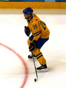 An ice hockey player dressed in a yellow and blue jersey holding his hockey stick in a ready position.