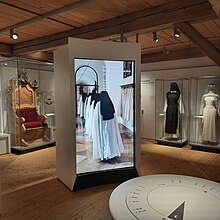 Auf einem großen Monitor Nonnen im Kloster, rechts dahinter Vitrinen mit Nonnentrachten, links eine Vitrine mit einem prunkvollen Äbtissinnensitz.