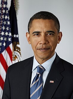 A portrait shot of a serious looking middle-aged African-American male (Barack Obama) looking straight ahead. He has short black hair, and is wearing a dark navy blazer with a blue striped tie over a light blue collared shirt. In the background are two flags hanging from separate flagpoles: an American flag, and one from the Executive Office of the President.