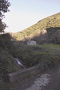 Petit pont sous lequel coule un filet d'eau. Au loin après un pré, l'église, dans l'ombre, puis un flanc de montagne ensoleillé.