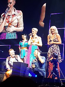 A group of five people - three women (pictured standing) and two men (pictured crouching) - standing on a stage performing a music act.