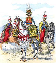Trois cavaliers équipés de costumes orientaux. Celui du centre dispose de deux grandes timbales sur son cheval.