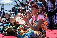 Une jeune femme assise en tailleur, jouant d'un tambourin. Elle est vêtue d'un grand pagne, et d'un tee-shirt rose.