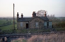 A photograph of the now demolished railway station at Constable Burton in Wensleydale, Yorkshire, England. A single railway line runs in front of the building. Two boarded-up windows are shown, as well as an open door which led into the old booking office. Four chimney stacks, some with large ornate Victorian chimneypots are on the roof. A telegraph pole stands to the left of the station.