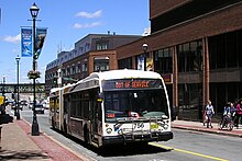 An articulated Halifax Transit bus on a city street. A brown brick, low-rise building is visible behind it on the left. On the right is the base of a high-rise building clad in the same kind of brick, with the signage of a CIBC bank branch visible on the ground floor.