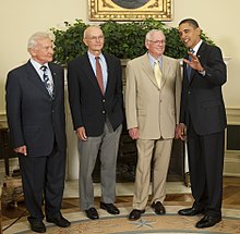 The astronauts are all elderly but standing straight. Aldrin wears a dark suit, Collins a dark sport coat and grey pants, and Armstrong a beige suit. The President is at the right. He wears a dark suit. He has medium-dark skin and is talking to Armstrong and raising his left hand. Armstrong is smiling.