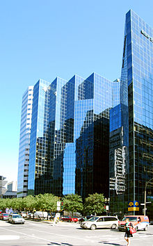 "Édifice à bureaux moderne aux parois vitrées bleu. Des arbres et une rue en bas de la photo et un ciel bleu clair encadre l'édifice