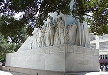 The rectangular base of a cenotaph. An angel is carved on one end. On the side are carvings of several men, shown wearing bucksin or 19th-century suits. Many hold guns or knives; at the far end, one operates a cannon.