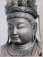 Portrait d'une statue en vue de trois quarts avec un visage plein et le lobe des oreilles allongé. Des petits visages sont situés sur le dessus de sa tête.