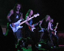 Four members of Iron Maiden are shown in concert. From left to right are a bass guitarist and then three electric guitarists. All members shown have long hair.