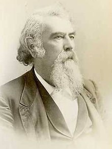 A man with white hair and a white beard and mustache, facing right. He is wearing a white shirt, black vest, and black jacket