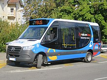 Photographie en couleurs d’un minibus bleu, blanc et noir de type Dietrich City 29, marqué J'ybus, et stationné à proximité de bâtiments résidentiels