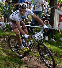 Homme sur un VTT portant un cuissard et un casque noirs et un maillot blanc à motifs bleus et noirs.