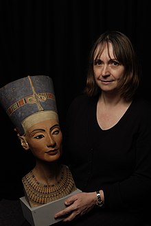 Une femme caucasienne aux cheveux chatains mi-long tenant dans ses mains le buste de Néfertiti.