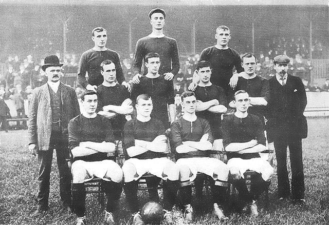 A black-and-white photo of a football team. The players are on three levels, with four on the bottom row, four on the middle row and three on the top row. The player sitting second from the left on the bottom row has a ball at his feet, and the player in the middle of the top row is wearing a flat cap.