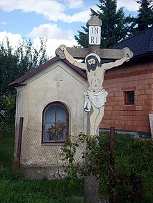 Uprostřed obrázku stojí výklenková kaplička ve špatném technickém stavu, před ní se tyčí krucifix s Ježíšem Kristem a nápisem INRI. Před kapličkou se nachází drátěný plot. Vzadu je vidět zahrada se zeleným trávníkem a část rozestavěného domu.