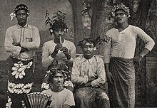 Photographie noir et blanc de cinq hommes vêtus du pagne traditionnel et portant des couronnes de fleurs. L'un joue de la flûte et un autre d'une sorte d'accordéon.