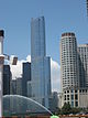 Distant ground-level view of a 100-story skyscraper with a blue, all-glass facade; the building has setbacks ant many levels, and a thin spire rises above its roofline.