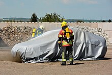 Ein brennendes Auto ist abgedeckt mit einer Brandbegrenzungsdecke, wenig Rauch ist zu sehen, zwei Feuerwehrleute mit Atemschutz beobachten die Situation.