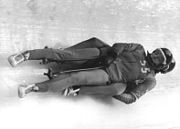 Two men equipped with dark jumpsuits, helmets and goggles slide down an ice track on a sled. One man is laying his back on the sled while the other is laying on top of his partner in the same position, and steers the vehicle.