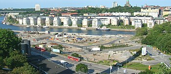 Området mellan Värmdövägen och Hammarby sjö. Den vänstra bilden är tagen den i juni 2005, och mitt i bilden syns Södermalms Trä:s brädgård, heminredningsbutiken Clark's Case och Norsk Hydros bensinmack. Till höger samma område i augusti 2009.