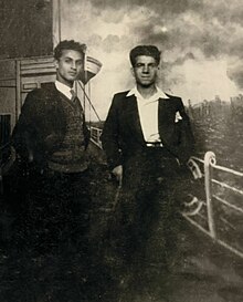 Photographie en noir et blanc de deux jeunes hommes debouts, vêtus de costumes et regardant l'objectif.