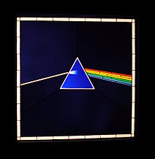 Triangle bleu rejoint depuis la gauche par un trait blanc et depuis la droite par un arc-en-ciel, le tout entouré par un carré blanc.