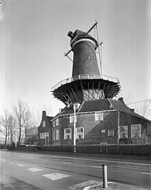Photographie en noir et blanc d'un moulin à vent sans ses ailes et d'une habitation longés par une route.