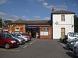 A brown-bricked building with a rectangular, dark blue sign reading "THEYDON BOIS STATION" in white letters all under a blue sky with white clouds