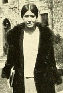 A young white woman, dark hair parted center and drawn back over ears to nape; she is standing outdoors, wearing a fur coat open in front, carrying several books in one hand