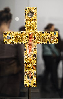 Ein reichhaltig mit Edelsteinen geschmücktes Goldkreuz in einer Vitrine, in der Mitte an zwei Positionen transparente Bergkristall-Behältnisse mit sichtbarem rotem Material im Inneren