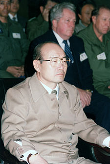 A portrait of an Asian man in his 40s wearing glasses and a beige wool jumper. He sits on a chair and narrows his eyes.