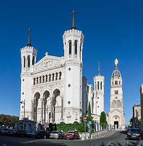 Photographie couleur montrant la basilique, au sommet de la colline, avec ses quatre tours et la statue de la Vierge, prise depuis le centre-ville de Lyon