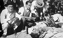 A black-and-white photograph of several men in early-20th century casual attire crouching down and inspecting a pile of dead bodies