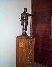 Bronze statuette of a man coaching hockey while wearing skates and holdindg a stick, mounted upon a tall wooden plinth with name plates for each recipient of the trophy.