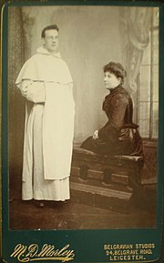 Photographie en noir et blanc ancienne d'un jeune moine debout, à côté duquel est assise une femme plus âgée.