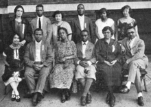 Twelve African-American adults, seven women and five men, posed outdoors for a group photograph, one row standing, one row seated.