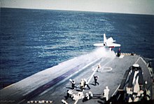 Colour photograph of a white military aircraft having just taken off from the flight deck of an aircraft carrier at sea