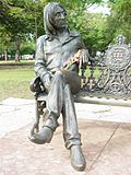 Statue of Lennon, bespectacled with long hair, on a park bench. There are red flowers in the statue's lap, and numerous trees are visible in the background.