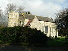 A flint church seen from the southwest. From the left are a round tower with an octagonal top, the west gable, the south porch, and the south wall of the nave with large windows