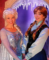 Deux femmes sont déguisées en personnage d'Elsa (robe bleue, perruque blonde) et d'Anna (robe noire et bleue, perruque rousse avec des mèches blanches).