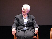 Sitzende Frau mit kurzen weißen Haaren, ihr Körper ist dem Fotografen zugewandt, sie blickt leicht lächelnd zu Seite. Sie trägt einen dunkelgrauen Hosenanzug.