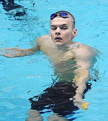 Hubert Kós im Schwimmbecken, der Kopf oberhalb des Wassers, der Körper im Wasser. Die Schwimmbrille hat Kós nach dem Rennen auf die Stirn hochgeschoben, in der Hand hält er die gelbe Badekappe.
